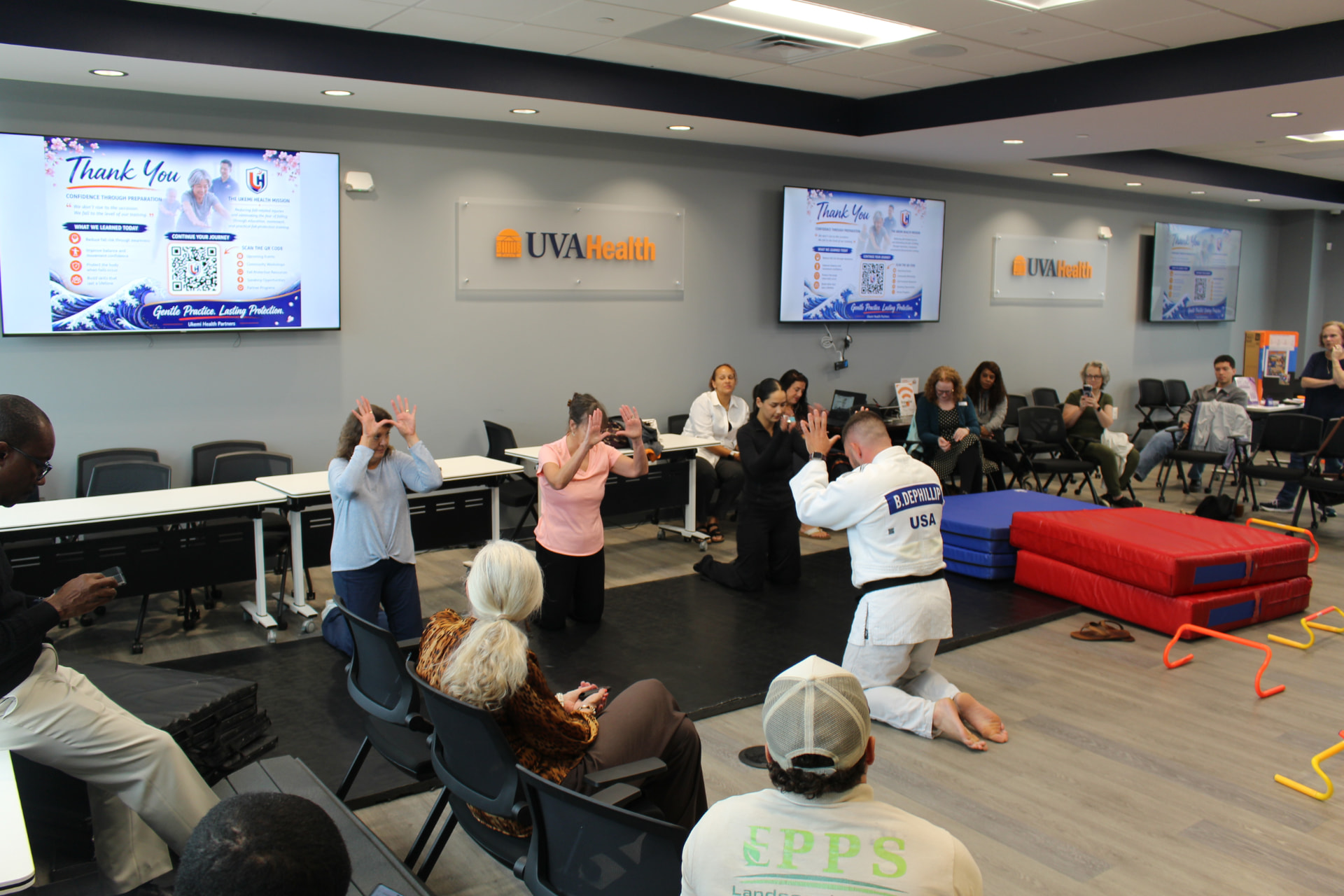 Bryan kneeling on a mat beside participants as they practice a protective arm position, while other attendees watch from their chairs